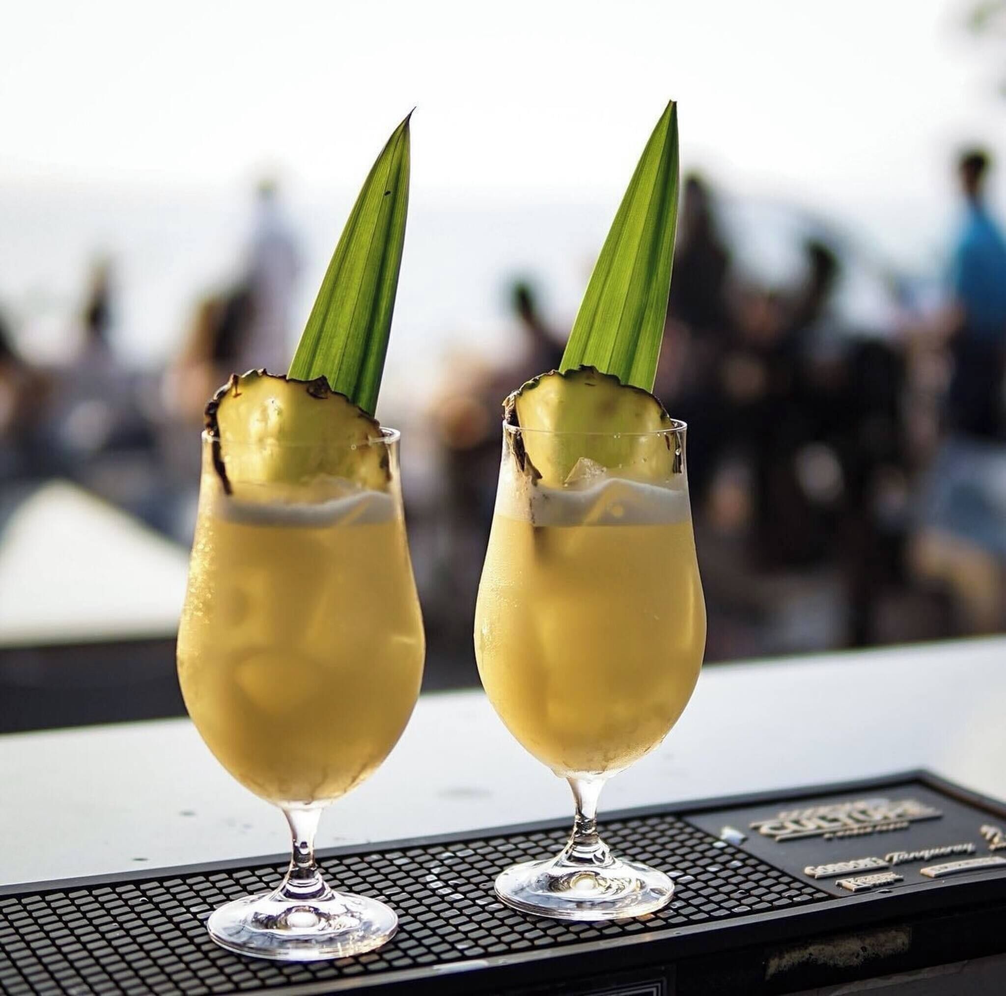 Two cocktails in stemmed glasses with a green leaf garnish, placed on a black tray with a blurred background of people.