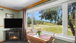 Una habitación con chimenea, televisión de pantalla plana, mesa de comedor con individuales y ventanas amplias que dan a un área con césped y árboles.