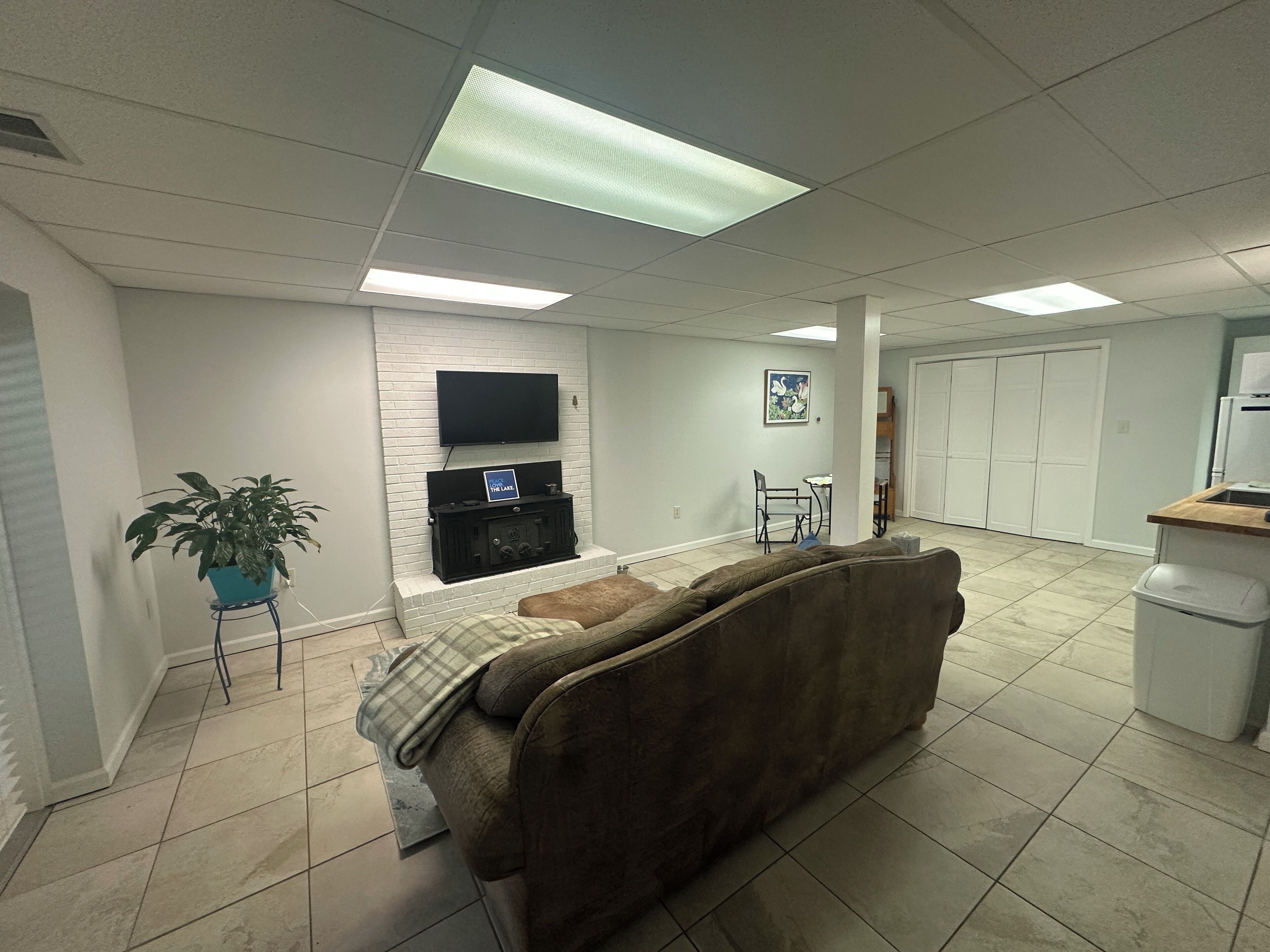 Een woonkamer met een bruine bank, een flatscreen televisie bevestigd aan een witte bakstenen muur, een klein tafeltje met stoelen en een potplant.