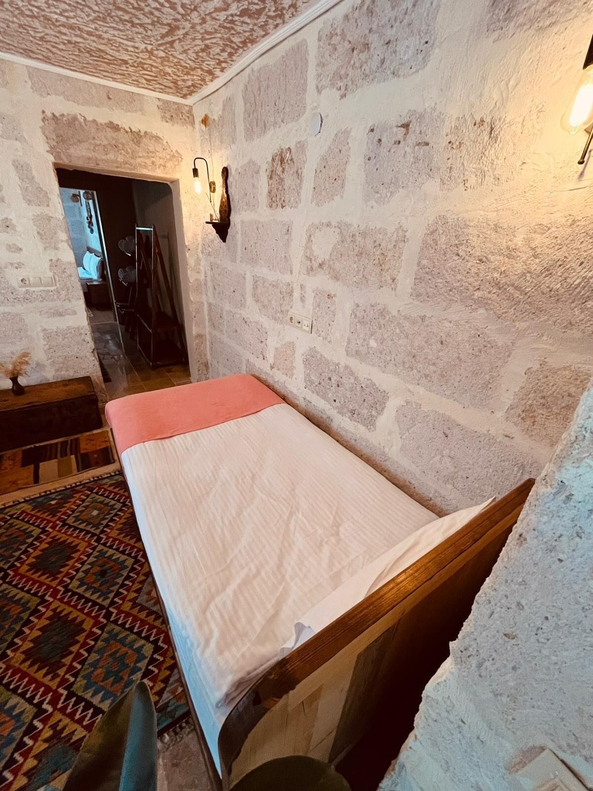 Una habitación con una cama individual, un tapete estampado, paredes de piedra y una pequeña mesa con sillas.