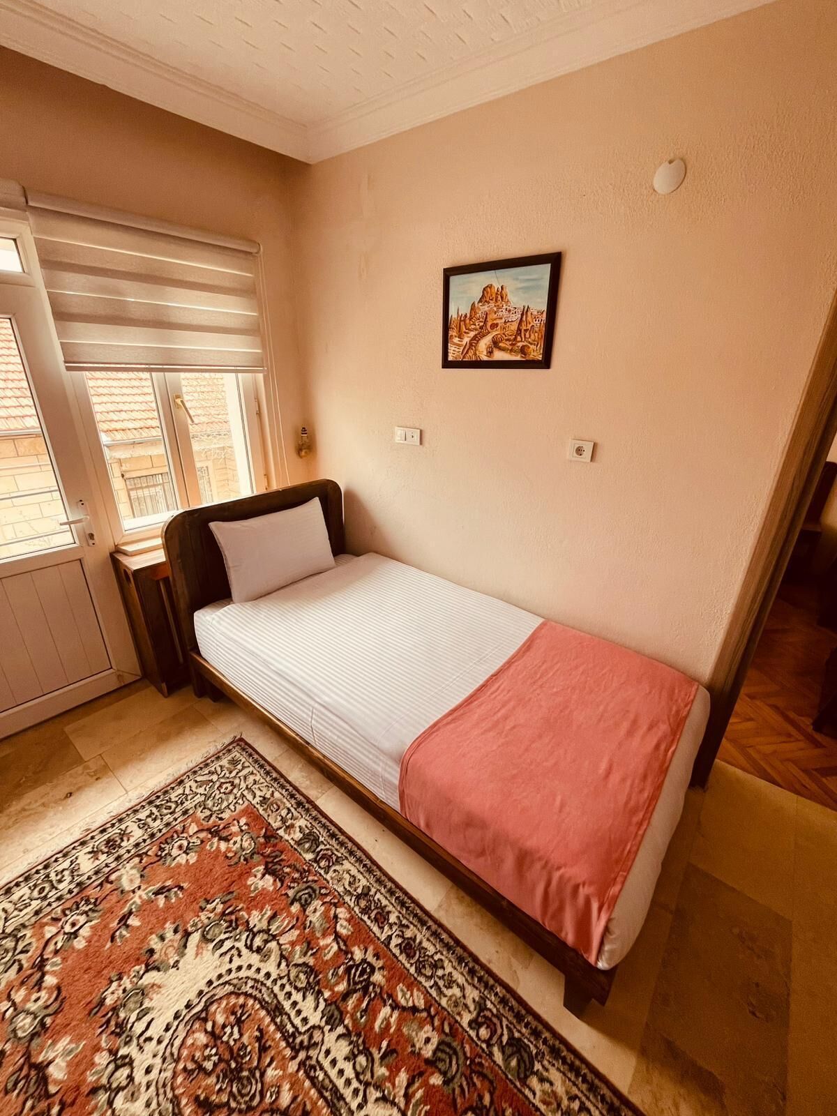 A single bed with a headboard, a patterned rug, a window with blinds, and a wall-mounted picture.