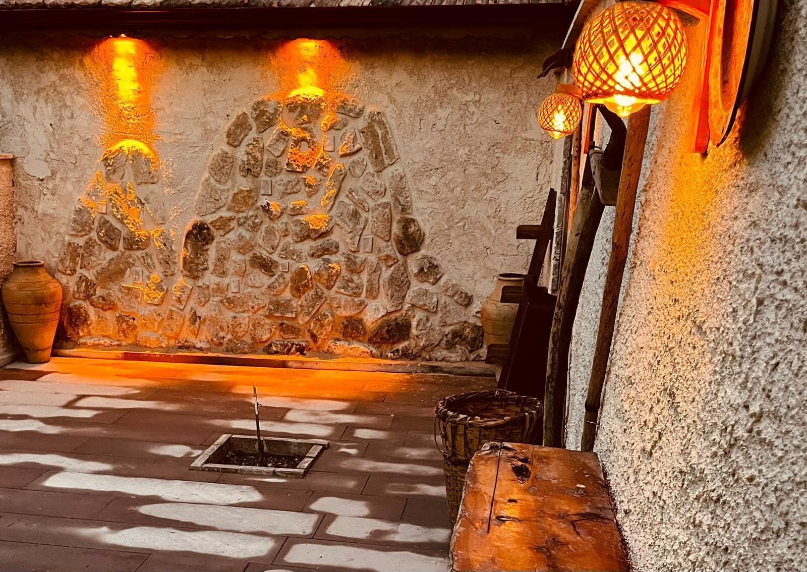 A stone-walled room with a central fire pit, a wooden bench, and a hanging lantern.