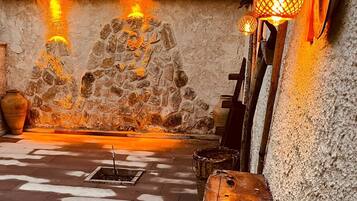 A stone-walled room with a central fire pit, a wooden bench, and a hanging lantern.