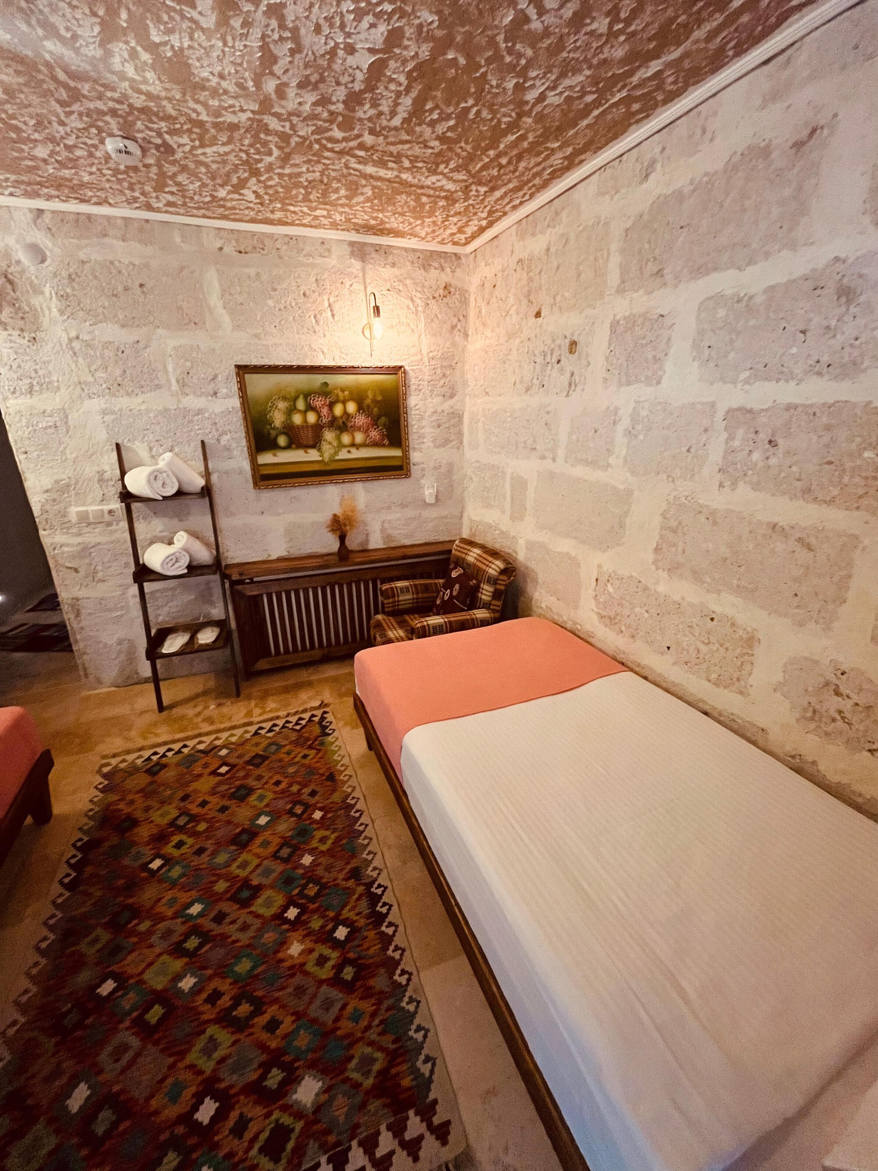 A room with a bed, a chair, a shelf with towels, a painting, and a patterned rug.