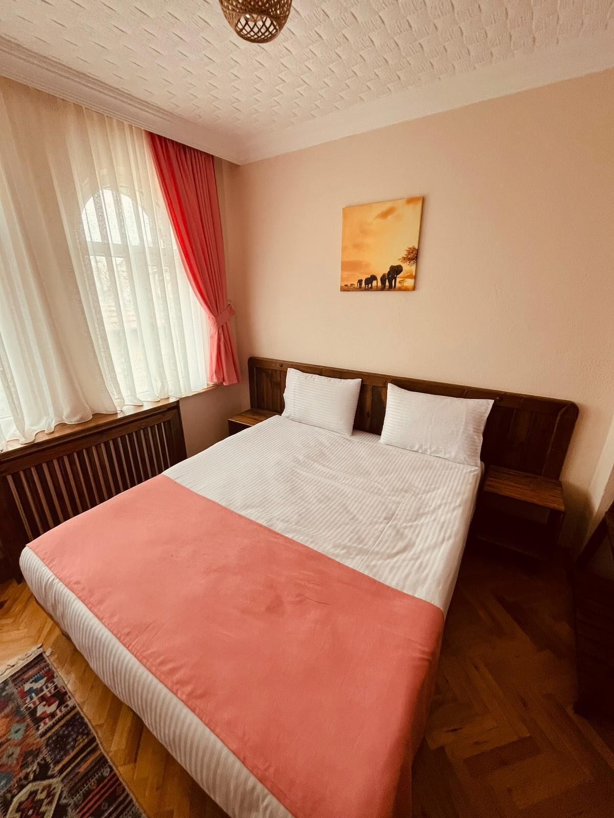 A bedroom with a bed, a window with curtains, a radiator, a wooden headboard, and a wall-mounted picture.