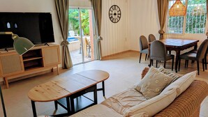 Un salon avec un téléviseur, des meubles en bois, un coin repas avec des chaises et une grande fenêtre avec des rideaux.