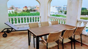 Une terrasse avec une table et des chaises, un transat et une vue sur les bâtiments et la verdure.
