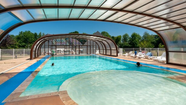 An indoor swimming pool with a glass ceiling, surrounded by lounge chairs and a covered area.