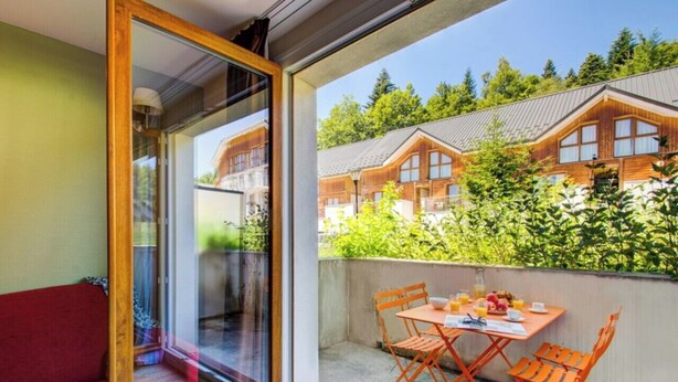 A balcony with a table and chairs, a red sofa, and a view of wooden buildings and greenery.
