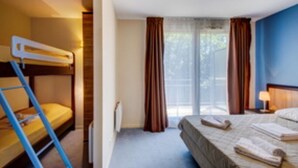 A hotel room with a bunk bed, a single bed, a window with curtains, and a bedside table with a lamp.