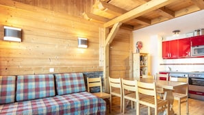 Ein gemütliches Blockhaushaus-Interieur mit Holzboden, einem karierten Sofa, einem Esstisch mit Stühlen und einer Küchenzeile mit roten Schränken und einer Mikrowelle.