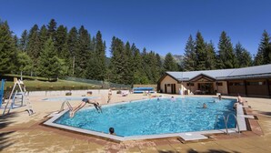 Ein Schwimmbecken unter blauem Himmel, umgeben von Bäumen und einem Gebäude mit einem Metall-Dach.