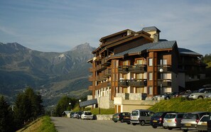 A multi-story building with balconies, a parking lot, and a mountainous backdrop. - Résidence Le Sappey Doucy 2P5S (La Léchère)