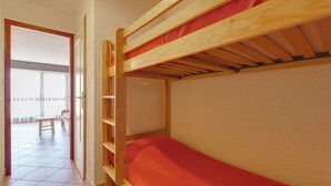 A bunk bed with a red mattress in a room with a tiled floor and a door leading to another room with a bench and a table.