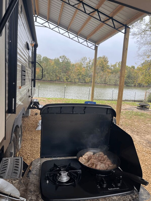 A portable gas stove with a pot of food, a covered area with a view of a lake, and a camper parked nearby.