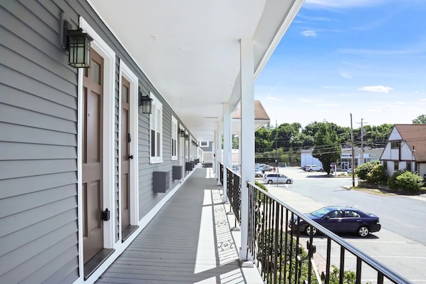 Une véranda couverte avec une balustrade, une rangée de portes et une vue sur une rue où sont garées des voitures.