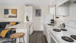 Una cucina compatta con mobili bianchi, un piano di lavoro bianco e un piano cottura integrato. C'è uno sgabello da bar e una piccola pianta sul piano.