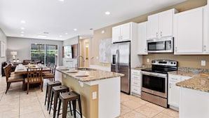 A modern kitchen with white cabinets, a stainless steel refrigerator, a microwave, and an oven. There is a dining area with a table and chairs. - Encore Swanky (Kissimmee)