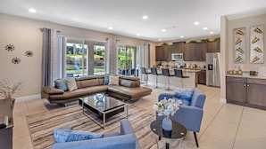 A modern living room with a sectional sofa, a coffee table, and a blue armchair. The kitchen features dark cabinets, stainless steel appliances, and a bar area. - Encore Swanky (Kissimmee)