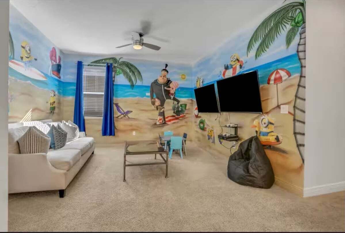 A room with a beach mural, a white sofa, a coffee table, a TV, and a ceiling fan.