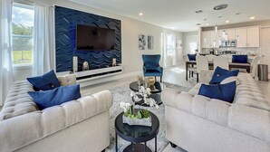 A modern living room with a large flat-screen TV, two sofas with blue cushions, a coffee table with decorative items, and a kitchen in the background. - Reunion Swanky (Kissimmee)