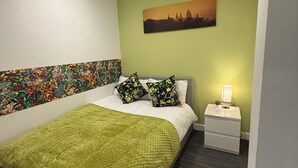 Un dormitorio con cama, mesita de noche, lámpara, pared verde y una pintura de paisaje urbano.