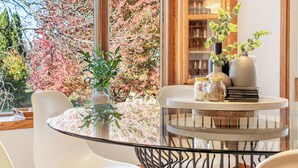 Un comedor moderno con una mesa redonda de cristal, sillas blancas y vistas a árboles otoñales a través de un ventanal grande.