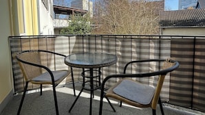 A balcony with a glass table and two wicker chairs, a metal railing, and a view of buildings and trees. - Charming apartment with terrace and parking space near the Rhine and Bonn city center (Bonn)