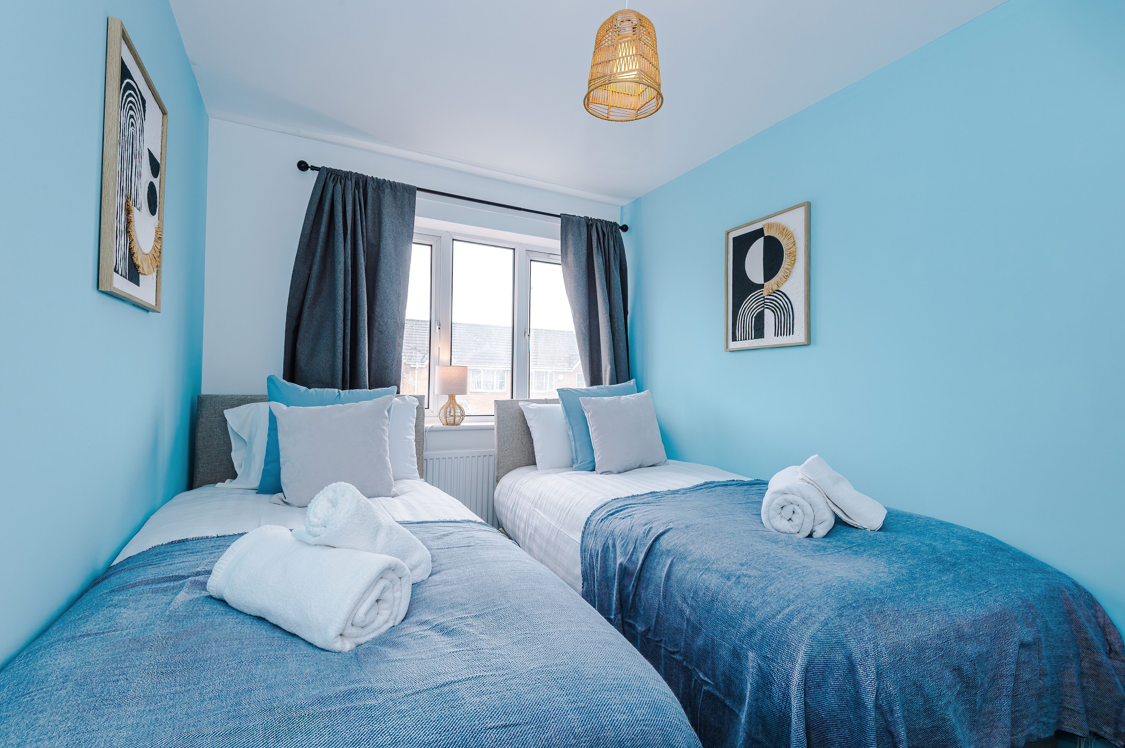 A hotel room with two beds, a window with curtains, a hanging light fixture, and artwork on the walls.