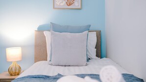 Ein ordentlich bezogenes Bett mit blau-weißem Kissen, ein Nachttisch aus Holz mit einer Lampe und ein gerahmtes Kunstwerk an der Wand.