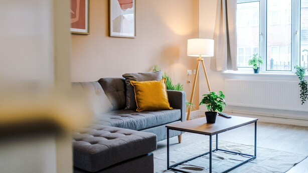 Ein modernes Wohnzimmer mit einer grauen Couch, einem kleinen Tisch, einer Stehlampe und einem Fenster mit Blick auf Gebäude.