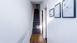 A narrow hallway with a wooden floor, white walls, and a staircase leading to a dark room. There are three framed informational posters on the wall.