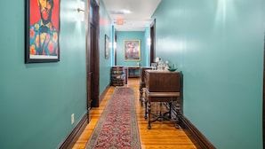 Un couloir aux murs de couleur turquoise, avec un plancher en bois, un tapis rouge à motifs, une table en bois sur laquelle se trouve un bol, et des œuvres d’art encadrées.