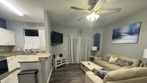 Une cuisine moderne avec des armoires blanches, un comptoir en granit et un four encastré. On y trouve un coin repas avec un canapé beige et une table basse. Un ventilateur de plafond et un tableau sont visibles sur les murs.