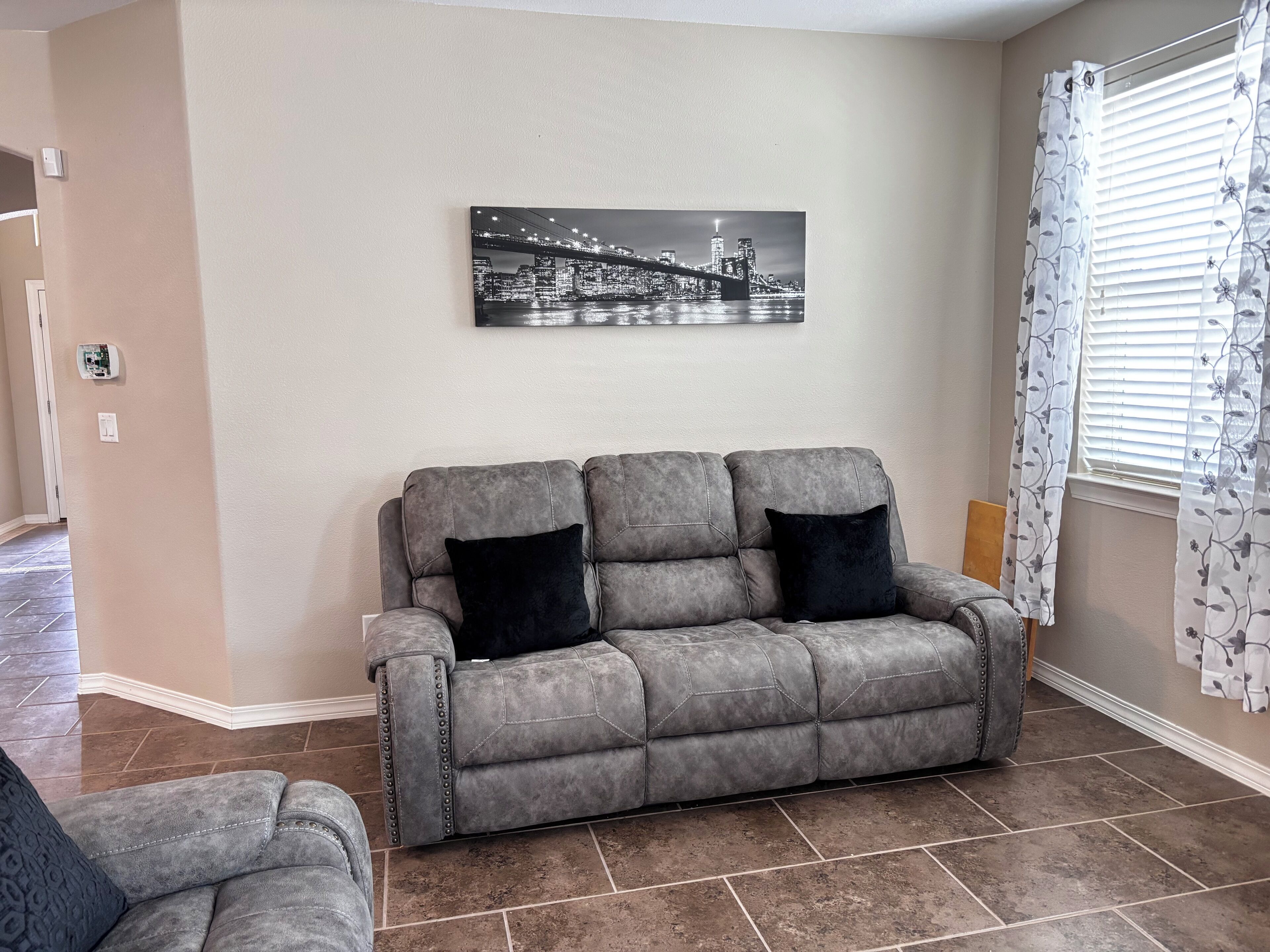 Un salon avec un canapé gris, deux coussins noirs, un tableau représentant un paysage urbain accroché au mur et une fenêtre ornée de rideaux à motifs floraux.