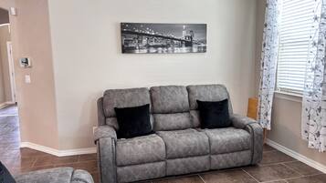 Un salon avec un canapé gris, deux coussins noirs, un tableau représentant un paysage urbain accroché au mur et une fenêtre ornée de rideaux à motifs floraux.
