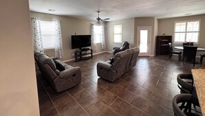 A spacious living room with a flat-screen TV, two recliner sofas, a ceiling fan, and a dining area with a table and chairs.