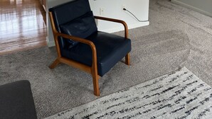 Ett vardagsrum med en mörkblå fåtölj, ett träbord, ett inramat fotografi på väggen och en matplats i bakgrunden.