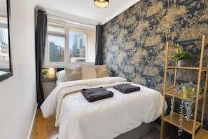 A bedroom with a bed, a window, a city view, a patterned wall, and a wooden shelf with plants.