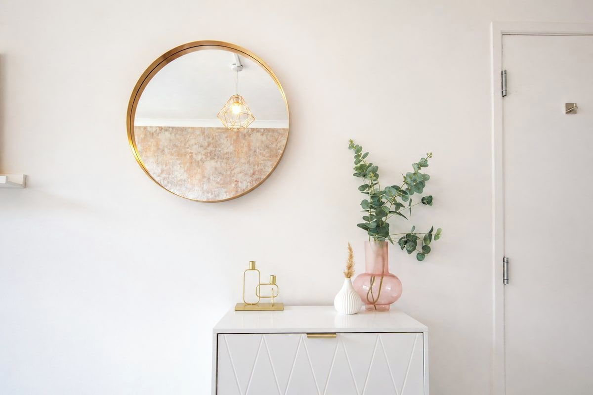 Un mueble blanco con un diseño geométrico, un espejo redondo con marco dorado y un jarrón con follaje verde.