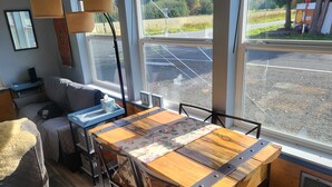 Un comedor con una mesa y sillas de madera, un sofá, una lámpara y vista a un estacionamiento a través de ventanas amplias.