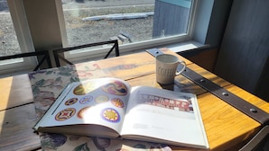 Un libro con diseños coloridos, una taza de café y un cuchillo sobre una mesa de madera junto a una ventana.