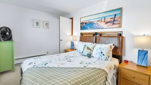 Hotelkamer met een bed, nachtkastje, groen kastje, wandventilator en een schilderij met een strandthema.