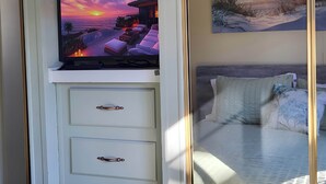 Un televisor mostrando una escena de atardecer, una cama con cabecero y un cuadro colgado en la pared.