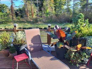 Eine Terrasse mit Liegestuhl und kleinem Tisch, umgeben von Kübelpflanzen und einem Holenzaun mit Blick auf einen Garten.