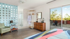 A bedroom with a large bed, a wardrobe, a seating area with a chair, a glass shower enclosure, and a view of the outdoors.