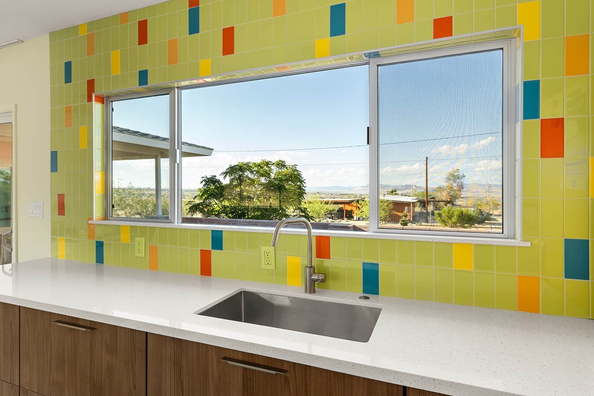 A kitchen with a large window, colorful tiles, and a stainless steel sink.
