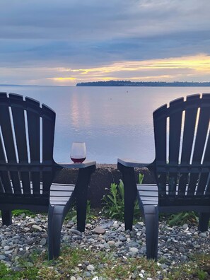 Two Adirondack chairs facing a body of water with a sunset in the background.