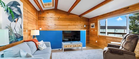 A cozy living room with a wooden interior, a blue accent wall, a fireplace, a sofa, a chair, and a large window offering a view.