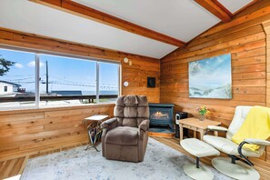 Une pièce chaleureuse aux murs en bois, avec une cheminée, un fauteuil inclinable et une vue sur l’extérieur.